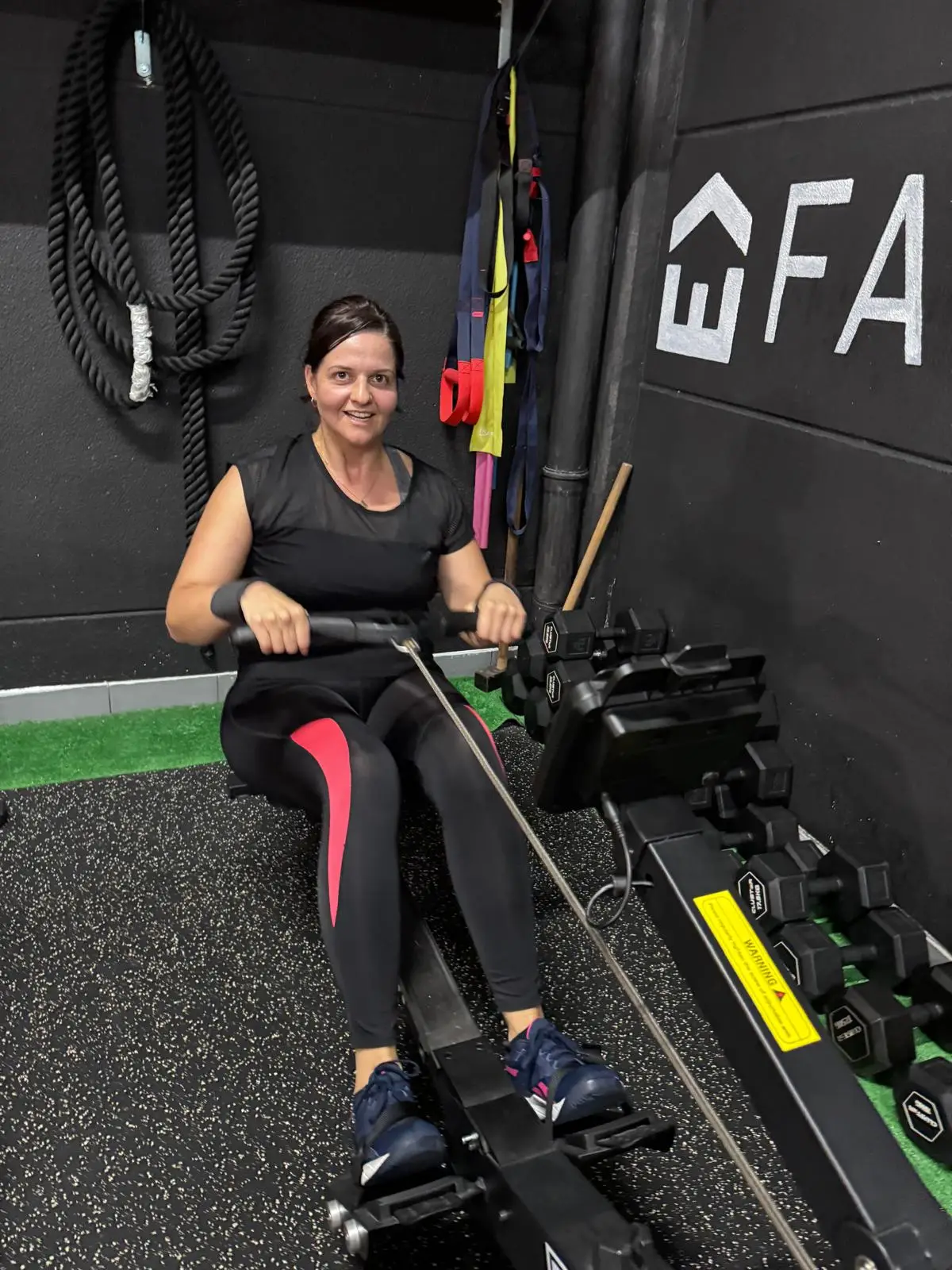 Atleta a remar no ergómetro, com as cordas e os halteres do estúdio atrás